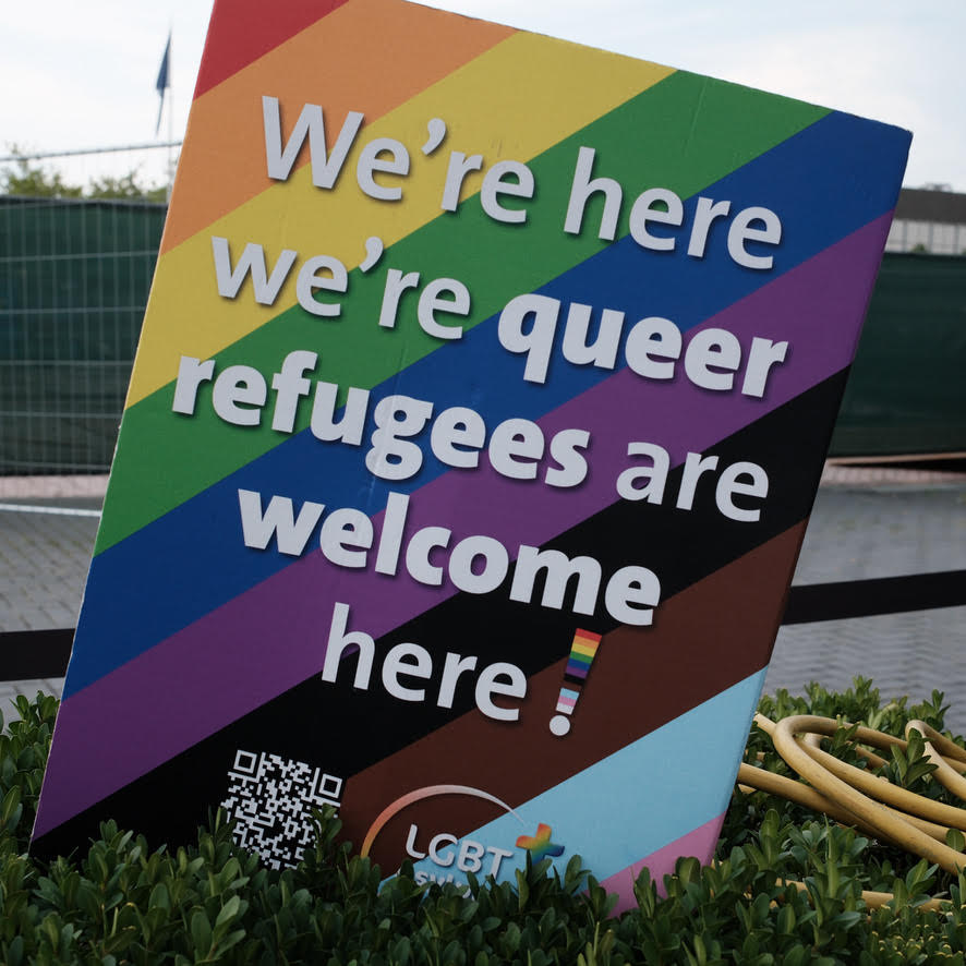 Bord met 'queer refugees are welcome here' op een regenboogvlag